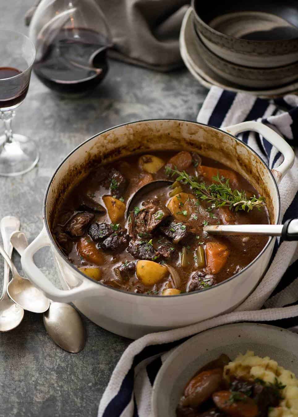 Beef Stew in a white casserole pot ready to be served into bowls beside it.